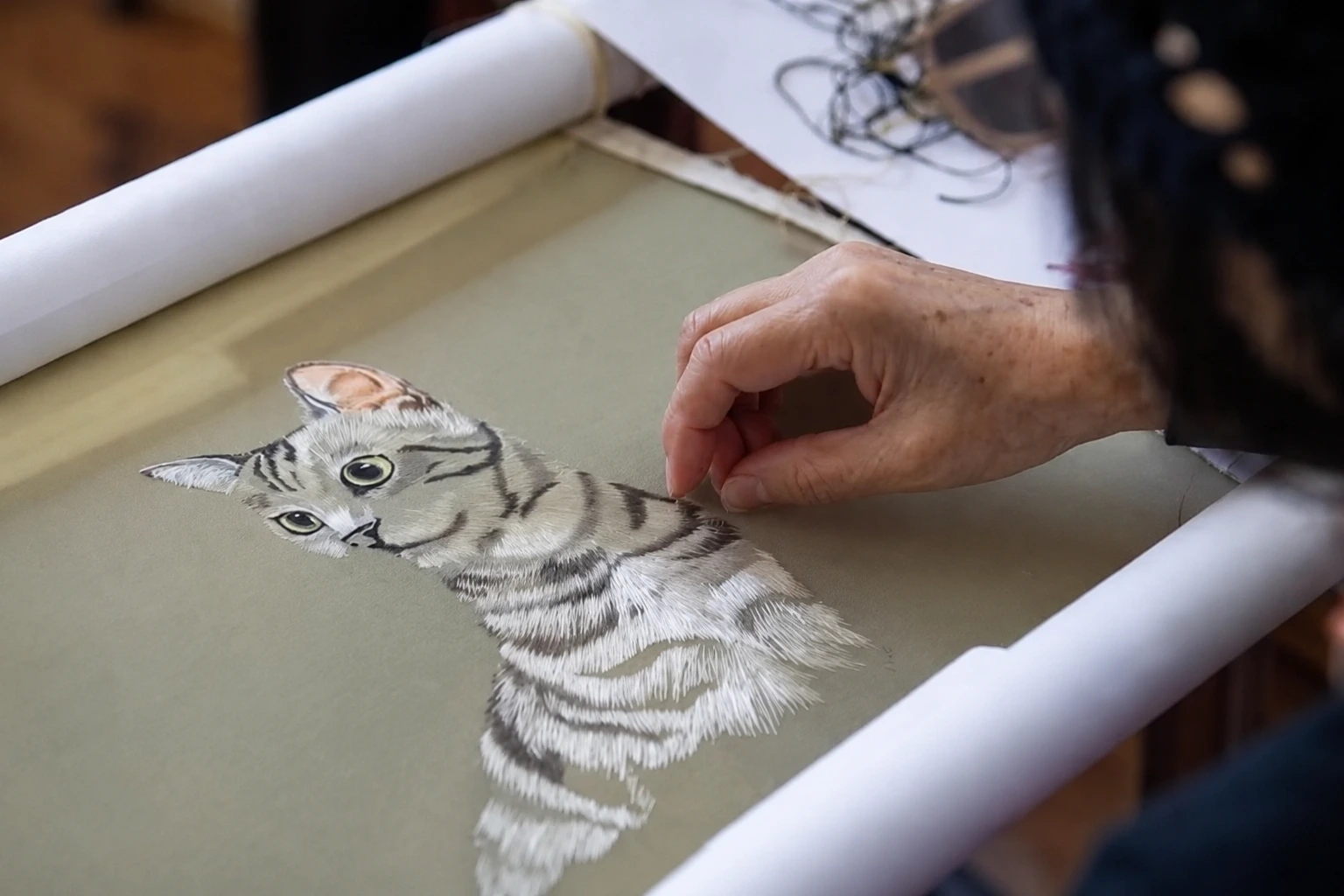 An elderly craftswoman stitching a detailed image of a playful cat onto silk with fine colored threads
