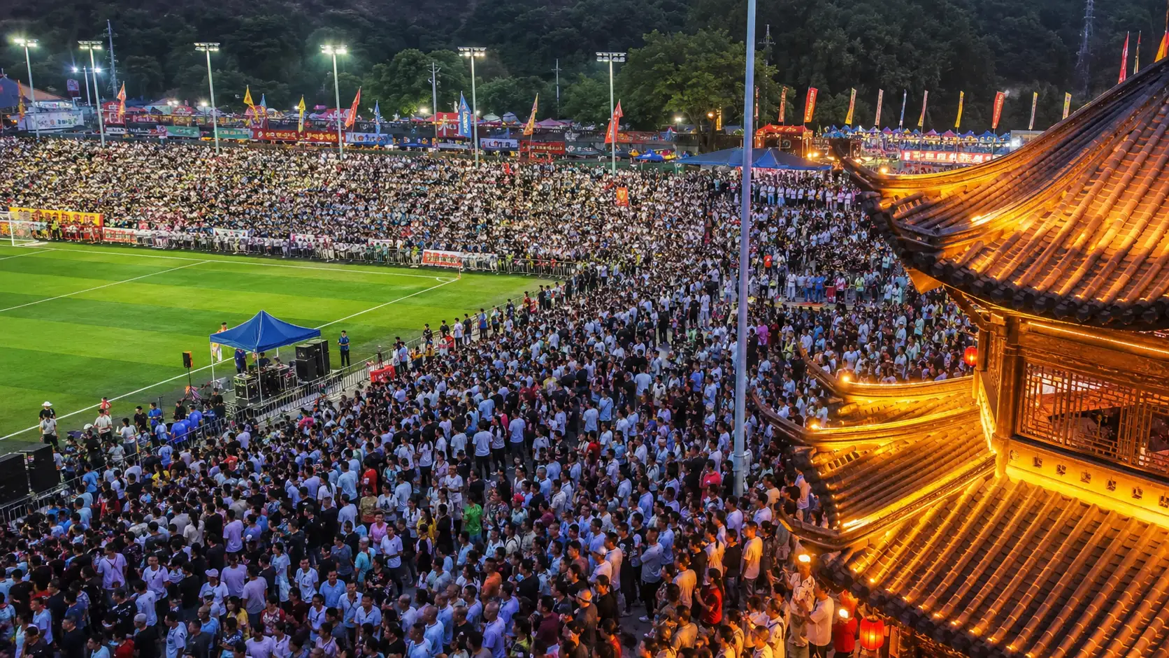Packed rural stadium at night with thousands of fans around a lit pitch