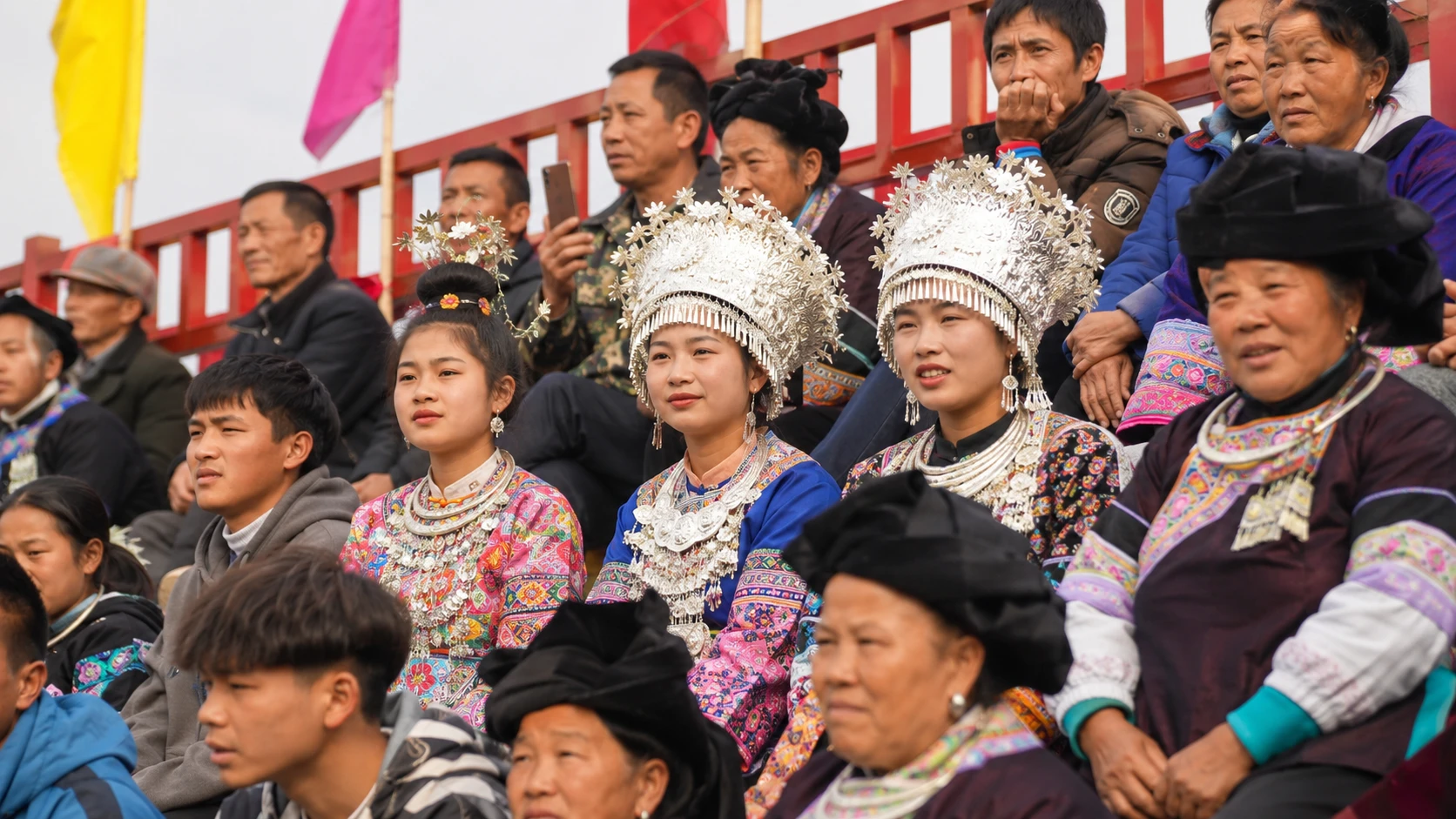 Women in silver headdresses and embroidered costumes watch from red bleachers