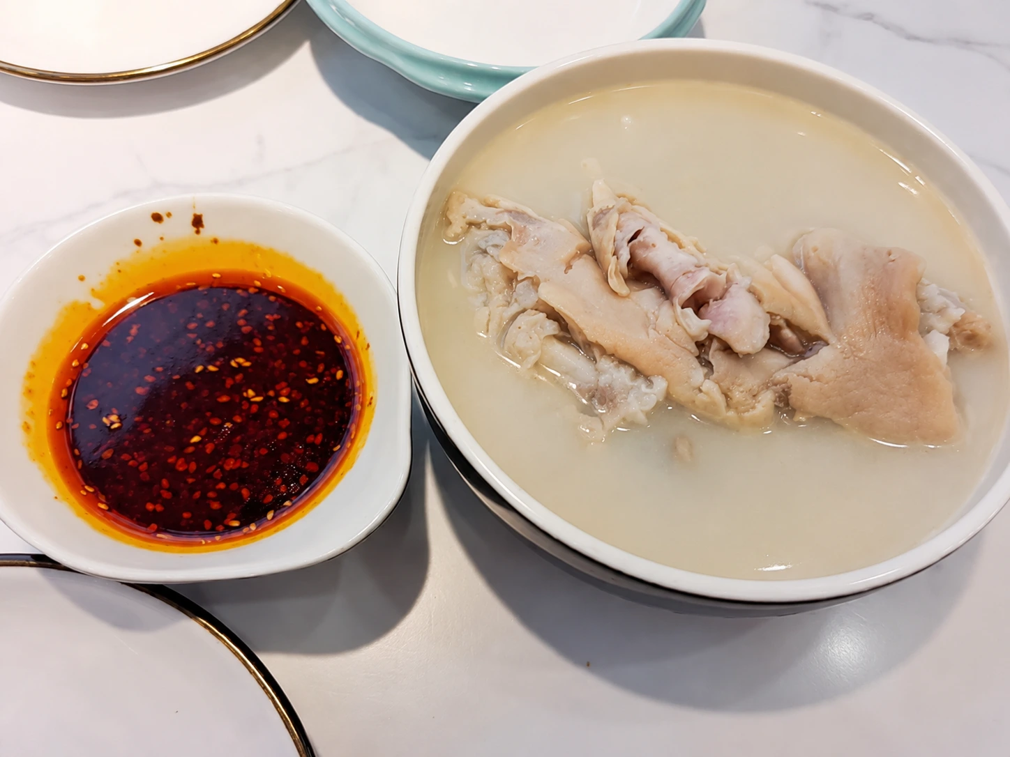 A small dish of chili-oil dipping sauce beside a bowl of mild pork hock soup