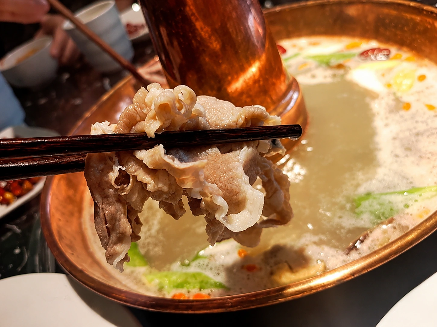 Paper-thin slices of raw lamb being lifted into a bubbling copper hot pot with chopsticks